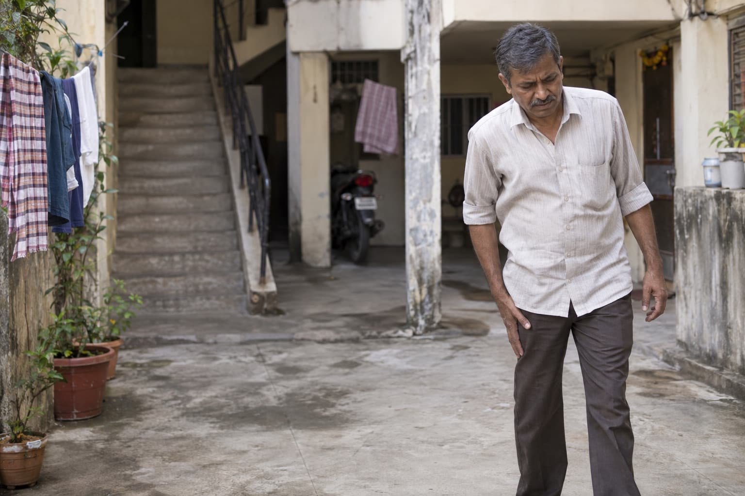 An Indian man pauses in a modest apartment courtyard with discomfort in one leg